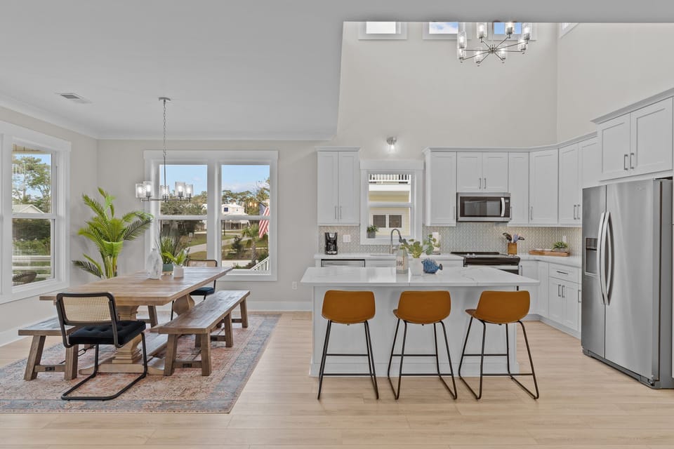 Bright dining space with wood table, bench, black chairs, chandelier, plants, large windows, porch views, patterned rug, and nearby modern kitchen.