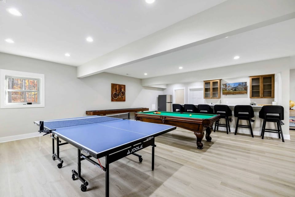 Entertainment room with a ping pong table, pool table, shuffleboard, and a bar area with seating, ideal for group activities and relaxation.