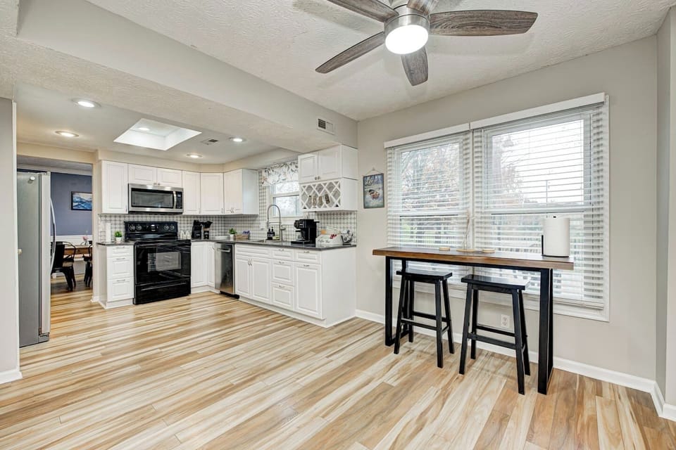 Bright, clean, modern, spacious kitchen with white cabinets, stainless steel appliances, & wood-patterned flooring. Adjacent to a dining nook are large windows, a high-top table, & stools. Enjoy breakfast with a beautiful and peaceful view of a lake