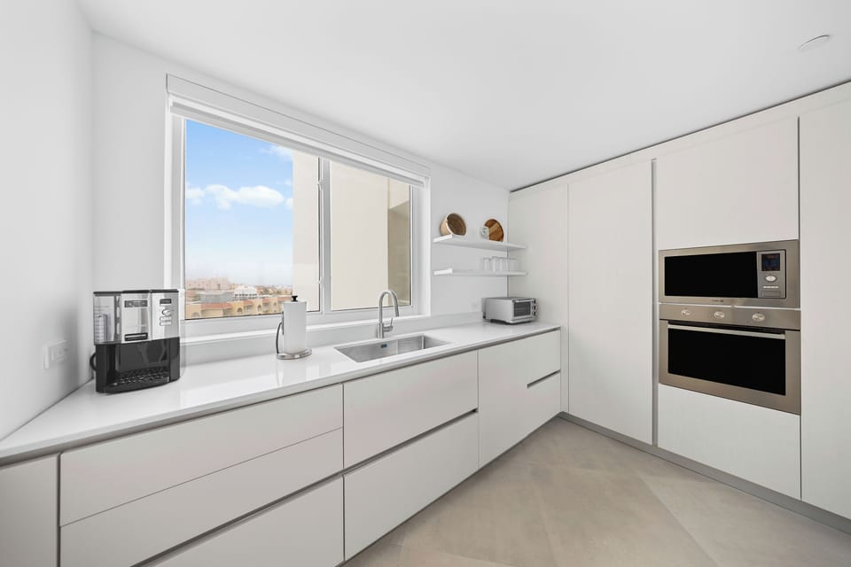 A sleek, modern kitchen awaits with gleaming white countertops and high-end appliances. Natural light streams through large windows framing city views, while you prepare meals in this bright, minimalist space.