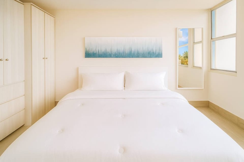 Unwind in this bright and peaceful king bedroom featuring crisp white linens, natural wood tones, and soothing ocean-inspired art. The soft light and simple design create a calm, hotel-style retreat perfect for relaxing after a day at the beach.