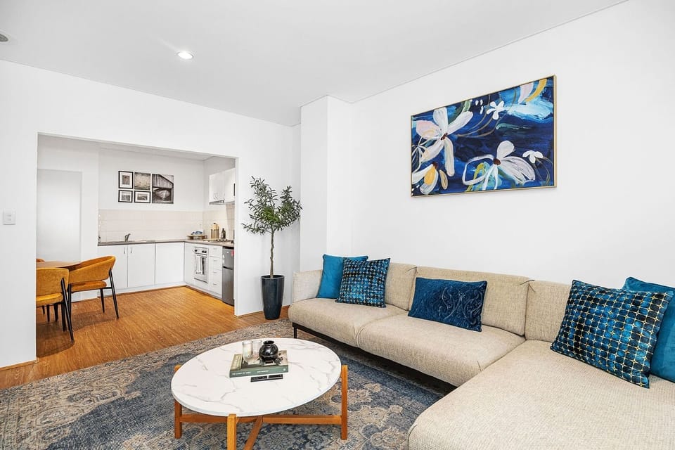 Enjoy an open-plan living area featuring a comfortable sofa and a colorful abstract artwork, seamlessly connected to a modern kitchen, creating a vibrant and inviting space.