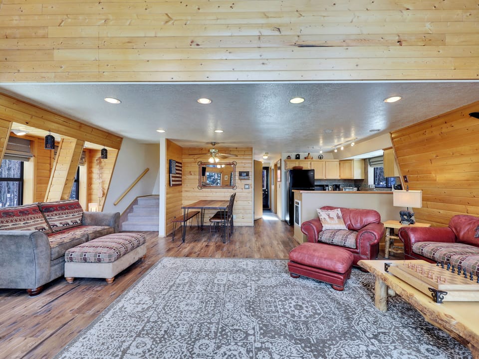 Great room featuring expansive tree views, access to an outdoor deck with a seating area, a cozy wood-burning fireplace, ceiling fans for added comfort, board games, a 43" Smart TV, and plush, comfortable seating.