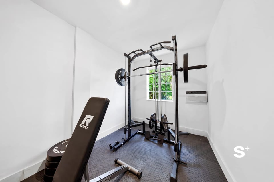 Focused gym area featuring a weight training bench with an attached barbell rack, weight plates, and a window for natural light.