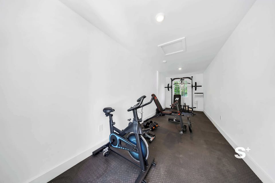 Private gym featuring an exercise bike in the foreground, a weight training rack, dumbbells, yoga mats, and dark rubber flooring.