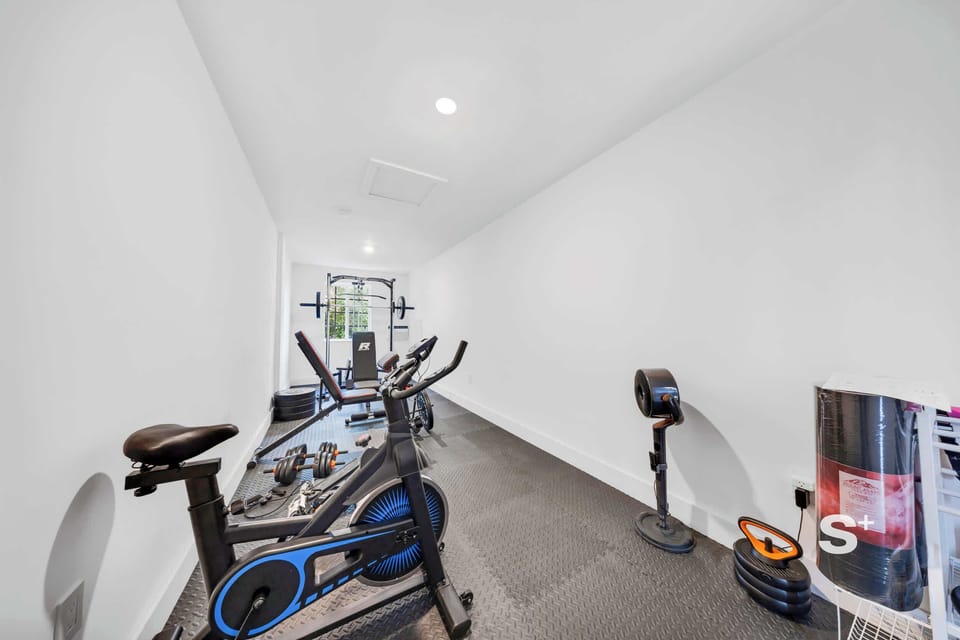Bright gym featuring an exercise bike in the foreground, a weight training rack, dumbbells, yoga mats, and dark rubber flooring.