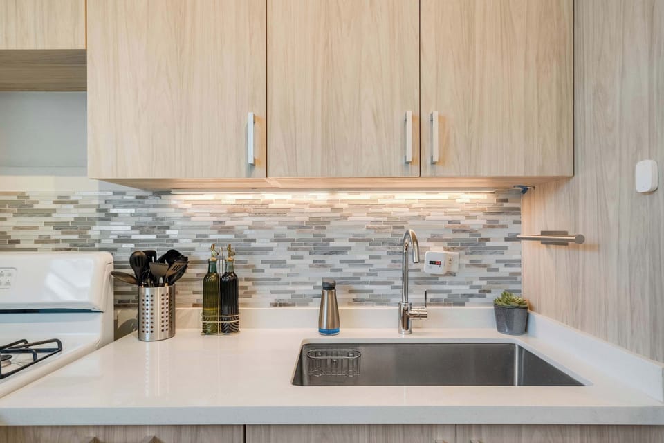 A narrow galley kitchen with light wood cabinets, a stainless steel gas stove, oven, and refrigerator. White countertops line both sides, with framed artwork on the wall and a wooden runner rug on the floor.