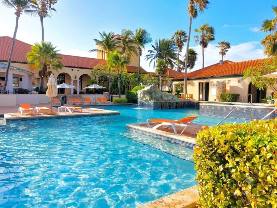A large swimming pool with a waterfall feature, orange lounge chairs, and palm trees.