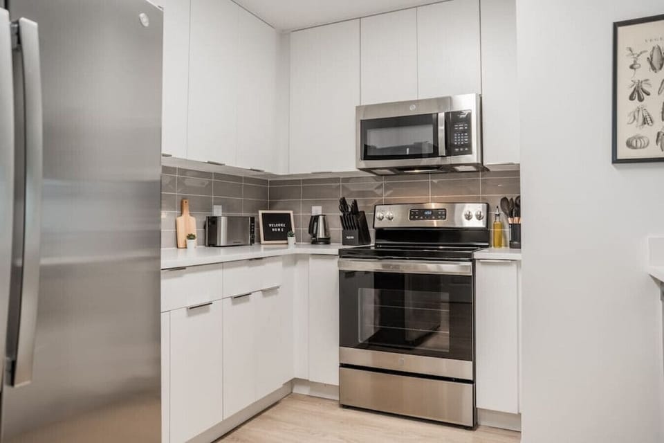 This modern kitchen features a spacious island with cabinetry, sleek white countertops, stainless steel appliances, open shelving, and stylish lighting.