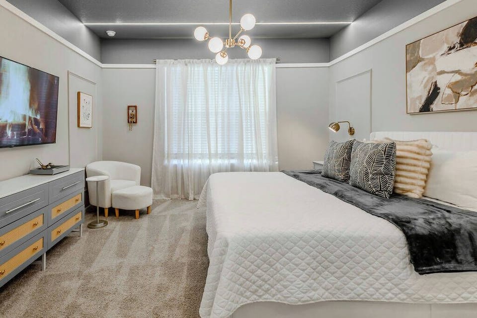 Experience a cozy ambiance in this stylish bedroom, offering a plush king-sized bed with soft linens and decorative pillows. The modern chandelier and neutral tones make it inviting, while a sleek dresser and comfy seating.