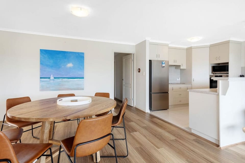 A modern, open-plan dining and kitchen area with a wooden dining table and brown leather chairs. The kitchen features a stainless steel fridge and built-in appliances. A large painting of a beach scene with a sailboat adds a touch of color and a coastal v