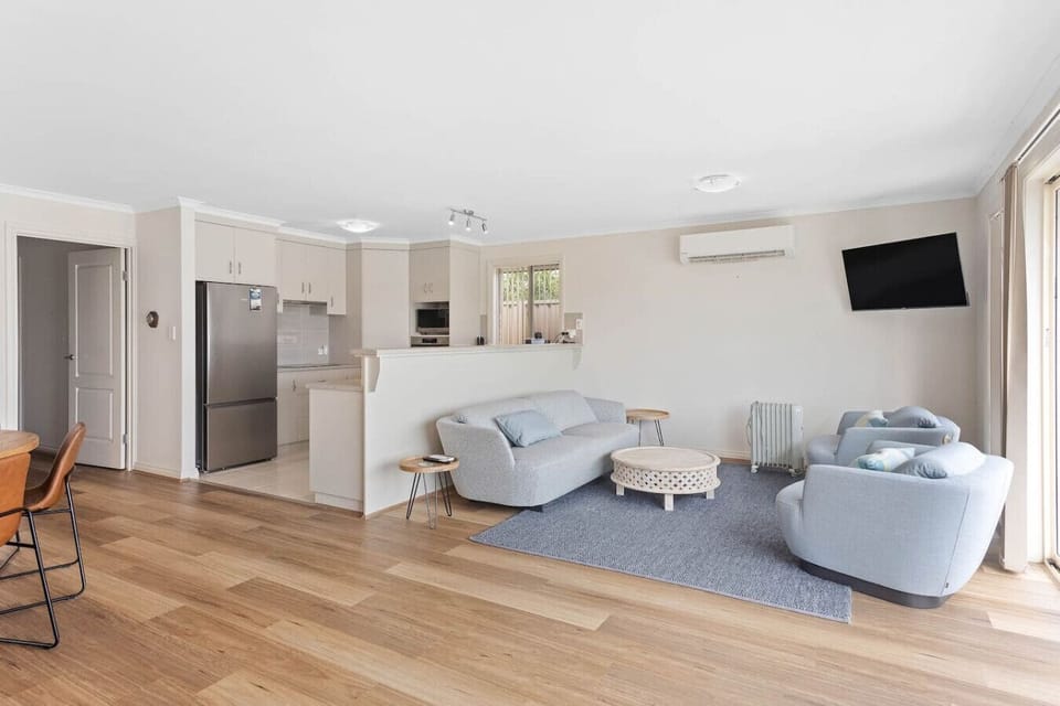 A modern, open-plan living area with light-colored walls and wooden flooring. It features a cozy seating arrangement with light gray sofas and a patterned round coffee table on a gray rug. The space includes a kitchen area with a stainless steel refrigera