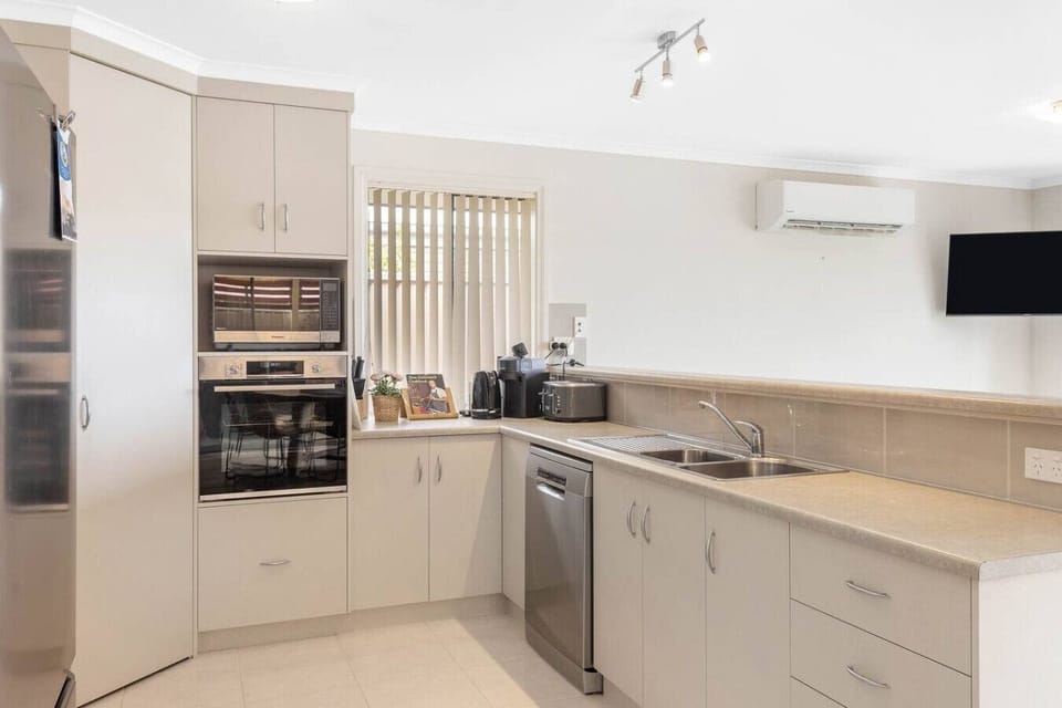 A modern kitchen with light-colored cabinets and a stainless steel dishwasher. It features a built-in oven, a microwave, a sink with a faucet, and a countertop with various small appliances. The kitchen also has a window with blinds and an air conditionin