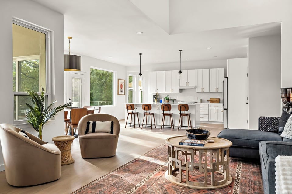 Stylish open-concept living space featuring a modern kitchen with white cabinetry, gold hardware, a large island with barstools, pendant lighting, a cozy living area with a plush sofa, accent chairs, a patterned rug, a round wooden coffee table, and large 