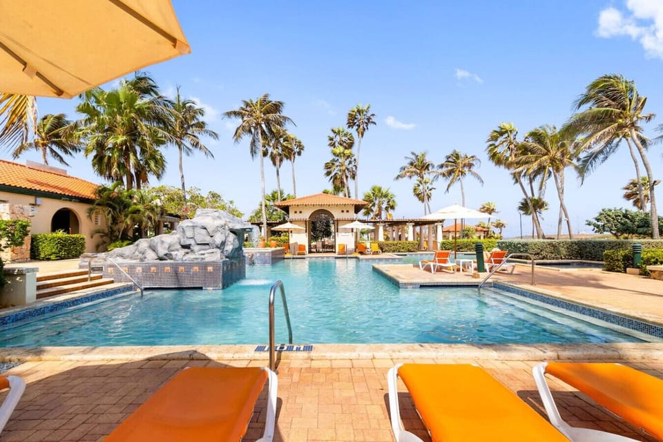 This vibrant pool area features a serene ambiance with inviting lounge chairs and shaded umbrellas, perfect for relaxation. The crystal-clear waters are accented by a decorative rock waterfall, enhancing the tropical vibe.