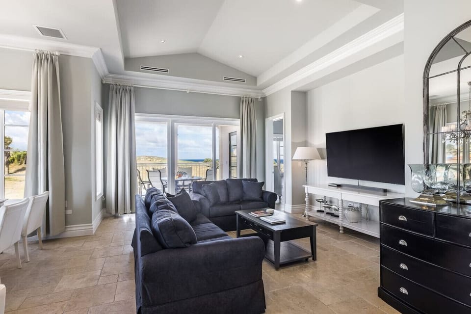 A bright, modern living room with navy couches, a sleek TV setup, and large windows showcasing stunning outdoor views. The airy space flows seamlessly to the dining area and terrace, blending comfort and style.