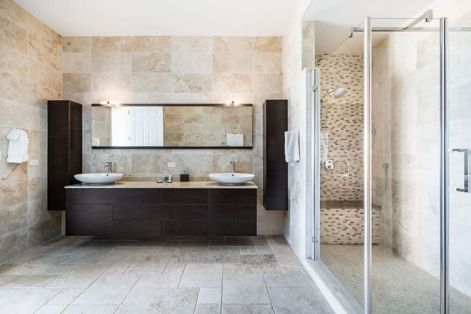 A modern bathroom with dual sinks, a large mirror, a glass-enclosed shower, and elegant natural stone finishes.