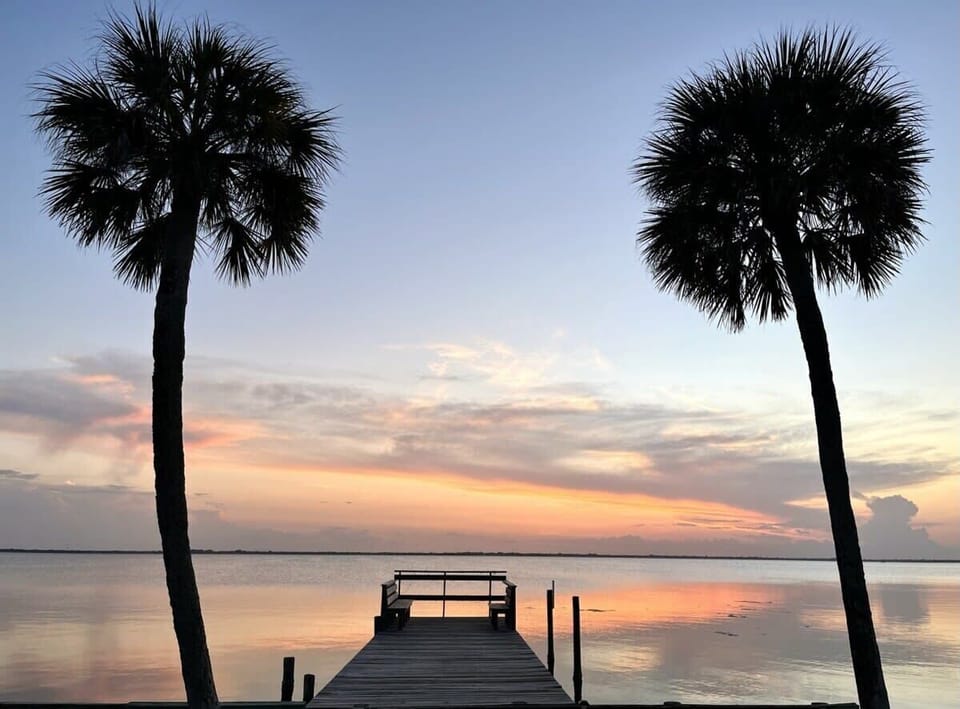 Relax on the wooden dock framed by palm trees with a serene sunset over calm waters.