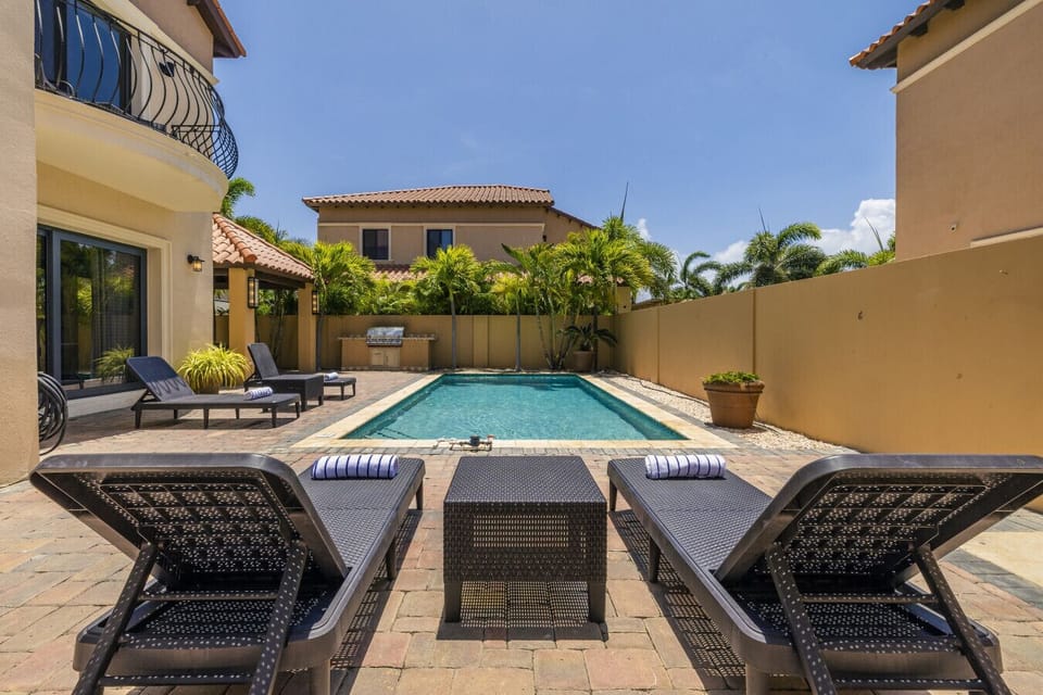 Relax by the serene poolside in this beautifully landscaped backyard, featuring stylish loungers, a cozy table, and tropical greenery. Perfect for enjoying sunny days and outdoor gatherings.