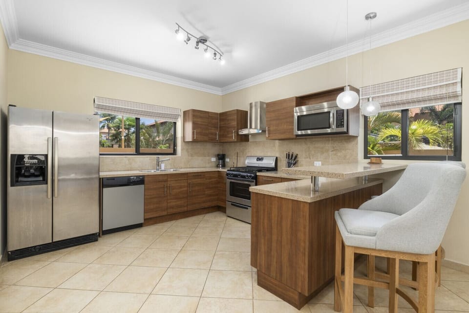 This modern kitchen features sleek wooden cabinetry, stainless steel appliances, and a spacious counter. Large windows provide plenty of natural light, making it a bright and inviting space for cooking and dining.