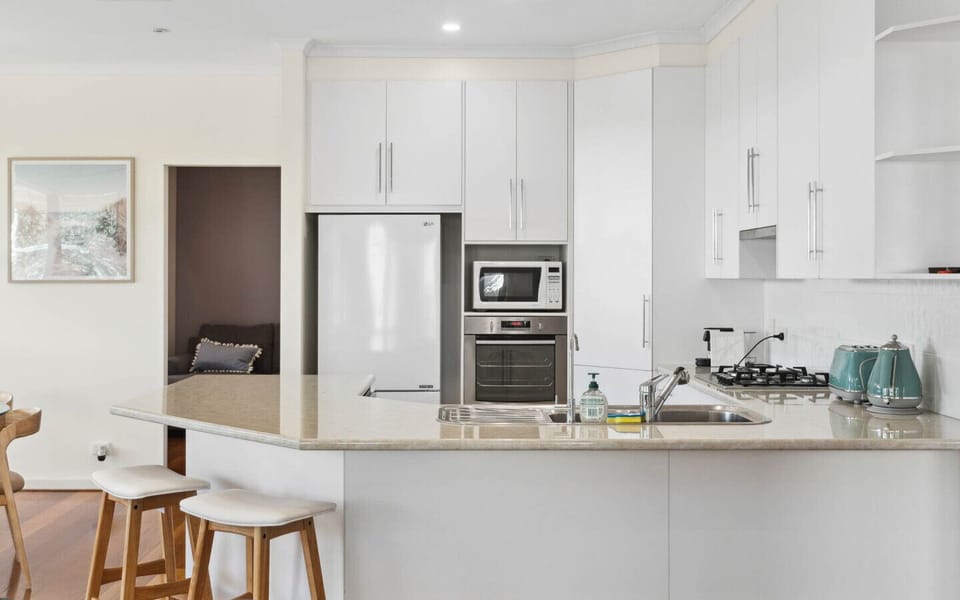 A modern kitchen with white cabinetry and a light-colored countertop. It features a microwave, an oven, a gas stove, and some teal kettles on the counter. There are also wooden stools in front of the counter, and part of a living area with a framed pictur