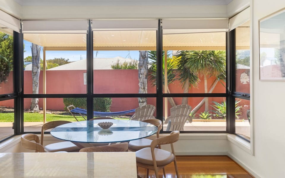 A cozy dining area with a glass-top round table and several chairs, situated by large windows that offer a view of a garden with palm trees and a hammock outside. The space has a bright and airy feel with wooden flooring and light-colored walls.