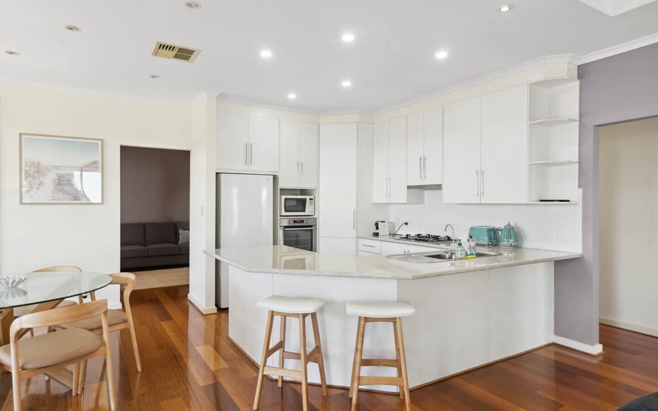 There are wooden stools at the kitchen island, and a glass dining table with wooden chairs is visible nearby. The kitchen is well-lit with recessed ceiling lights, and in the background, there is a glimpse of a living room area with a sofa. The floor is m