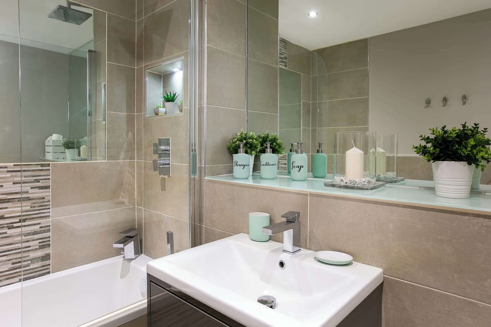Modern bathroom featuring beige tiles, glass shower screen, white fixtures, decorative mosaic strip, and mint green accessories with potted plants.