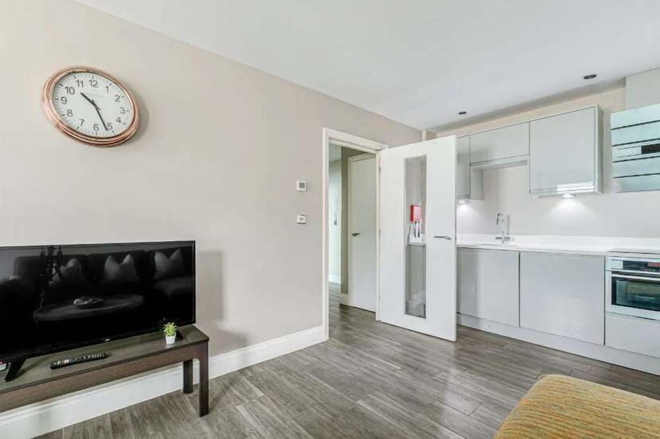 Open-plan living area with flat-screen TV, modern white kitchen, gray flooring, and minimalist decor.