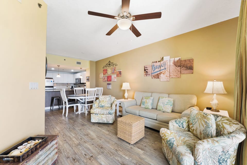 Relax in your bright, coastal-themed living space with comfortable seating, ceiling fan, and open concept flow to the kitchen.