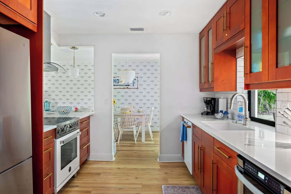 This image captures the full length of the kitchen, showcasing rich wooden cabinets and modern appliances. A view to the dining area beyond suggests an open-concept design, with natural light filtering in through large windows. 