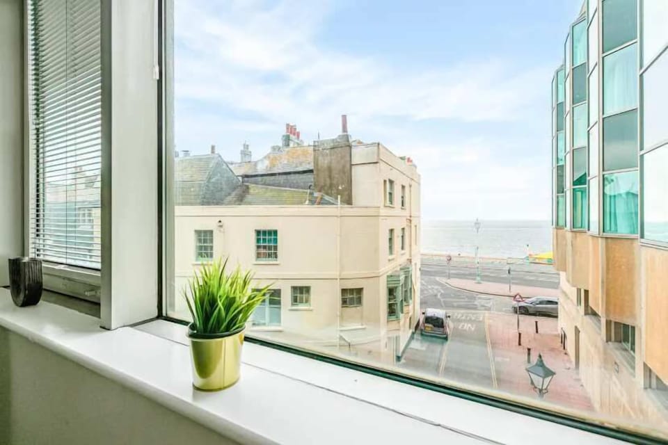 City view from apartment window showing historic buildings, seafront promenade, and modern glass architecture, with decorative plant on windowsill.