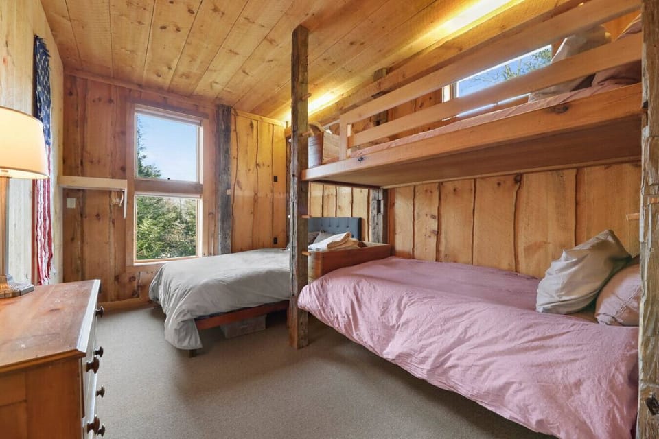 A warm, wood-paneled bedroom featuring a twin-over-twin bunk bed and a separate queen bed, ideal for kids or extra guests, with a large window bringing in natural light.