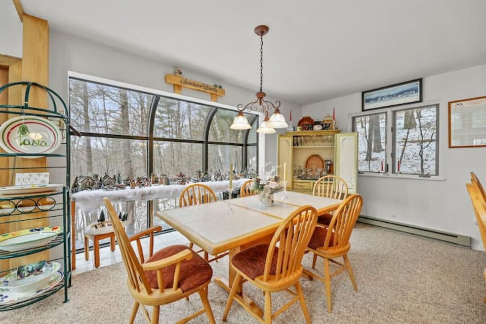 Cozy dining space with a wooden table and six chairs, surrounded by large windows that offer serene forest views, complemented by a vintage hutch and warm natural light.