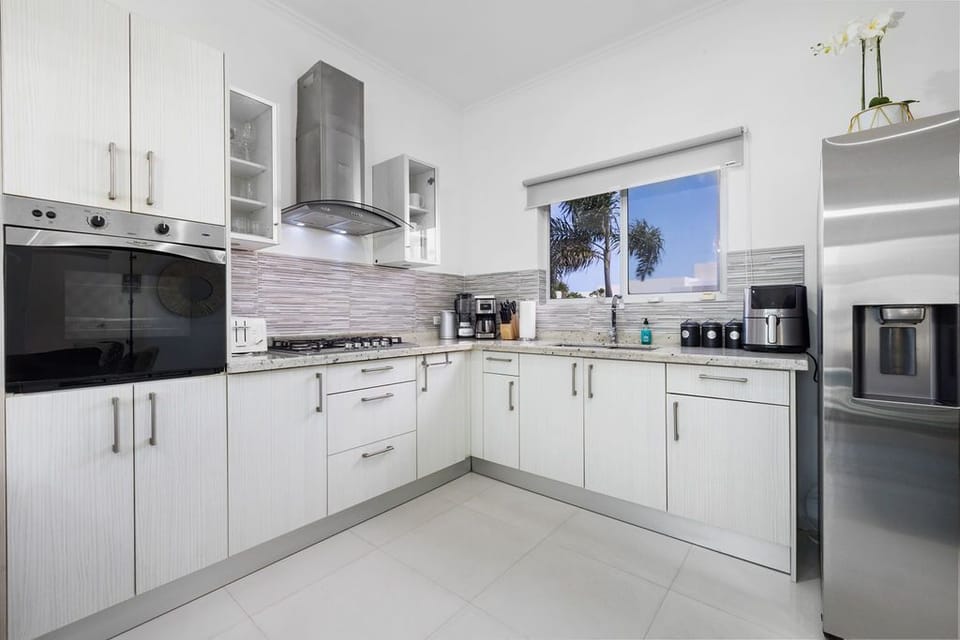 Your fully-equipped kitchen features modern appliances, ample counter space, and crisp white cabinetry bathed in natural light from the sunny window overlooking swaying palms outside.