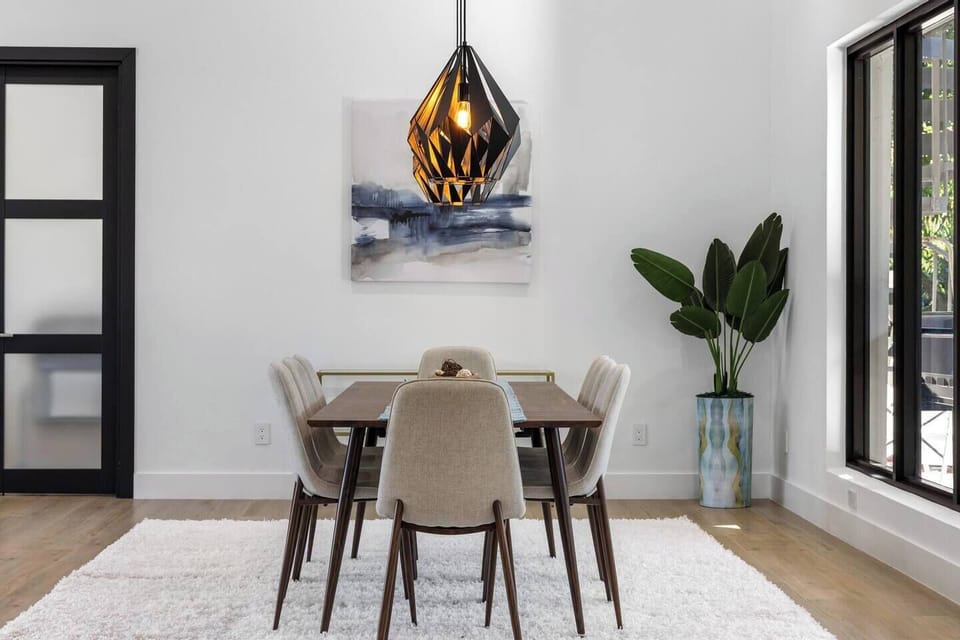 Bright dining area - wood table seating six, sculptural pendant light, and floor-to-ceiling black-frame windows.