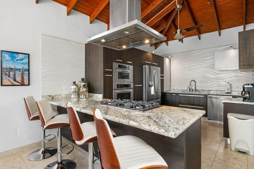 A sleek, modern kitchen with a granite countertop island, complete with bar stools, a built-in stove, and a stainless steel range hood. The open design features dark cabinetry, a vaulted wooden ceiling, and premium appliances for  sophisticated look.