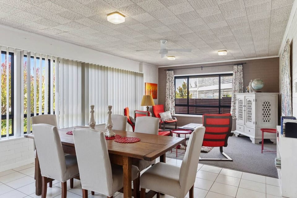 A bright and airy living and dining area with a wooden dining table surrounded by light-colored chairs. The space also features a seating area with red and white furniture, a ceiling fan, and large windows with vertical blinds that let in natural light.