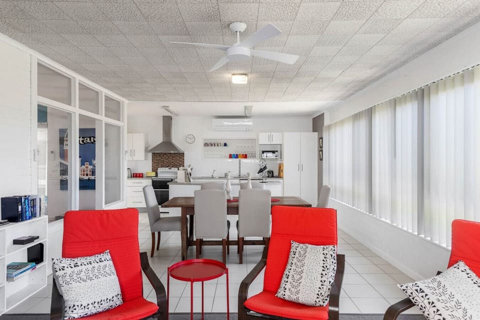 The room includes a kitchen area with a stove and cabinets, a dining table with chairs, and a cozy seating area with bright red chairs adorned with patterned cushions. A white ceiling fan adds to the comfortable and inviting atmosphere.