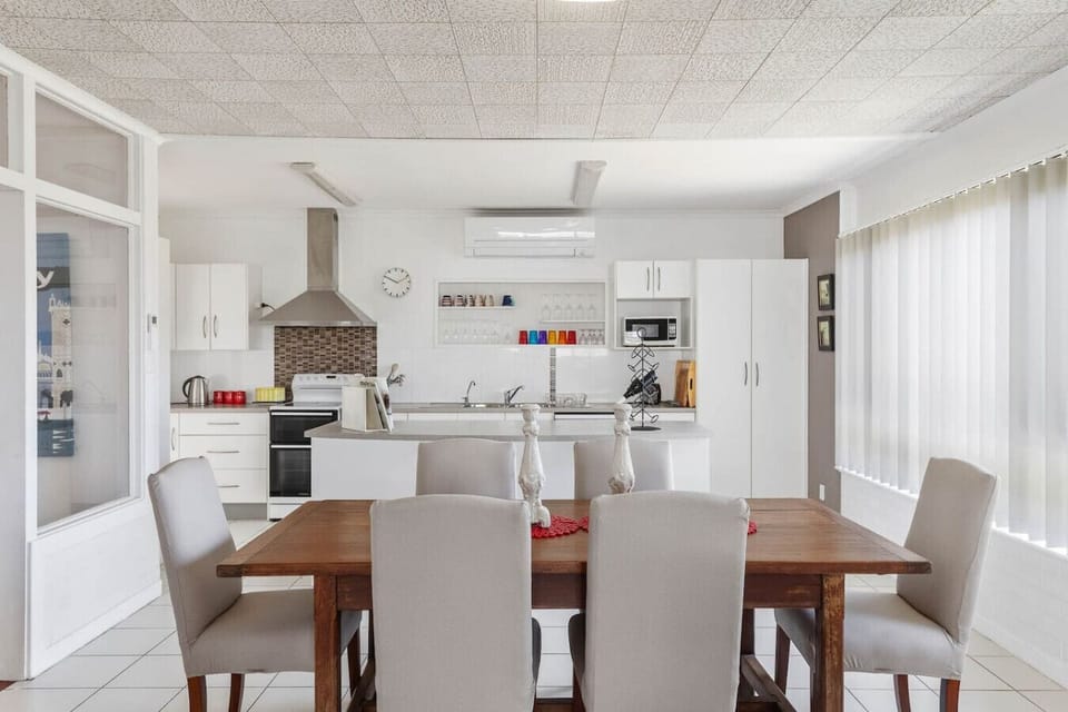 The kitchen features white cabinets and appliances, including a stove and a microwave, along with a tiled backsplash. Adjacent to the kitchen is a wooden dining table surrounded by light-colored chairs, creating a cozy dining space.