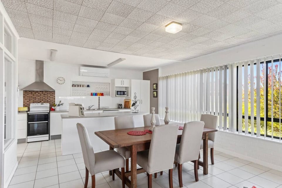 A bright and airy kitchen and dining area with a modern design. It features white cabinetry, a stainless steel stove, a wooden dining table with light-colored chairs, and large windows with vertical blinds that let in natural light and offer a view of som