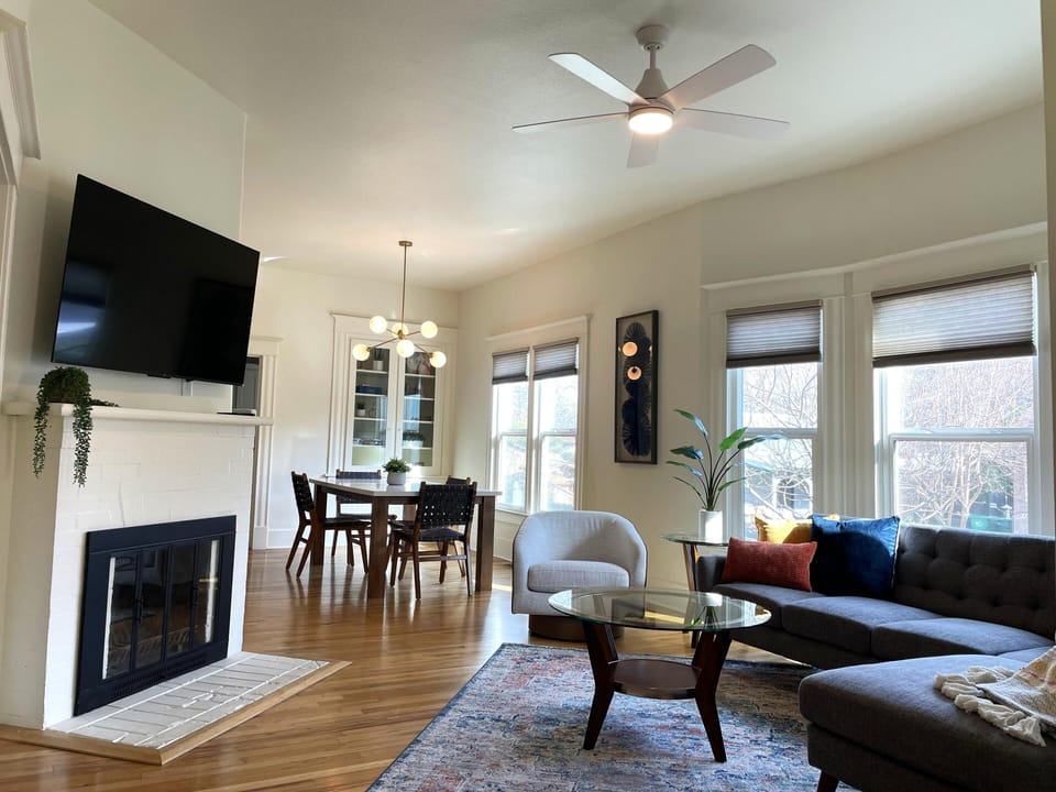 Spacious living room has TV with streaming services, ceiling fan, window shades.