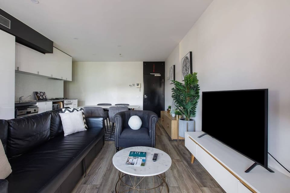 Relax in a light-filled living area of apartment one with timber floors, plush seating, and a large TV, all enhanced by a touch of greenery. Step directly onto the balcony to enjoy the fresh air and Melbourne skyline.