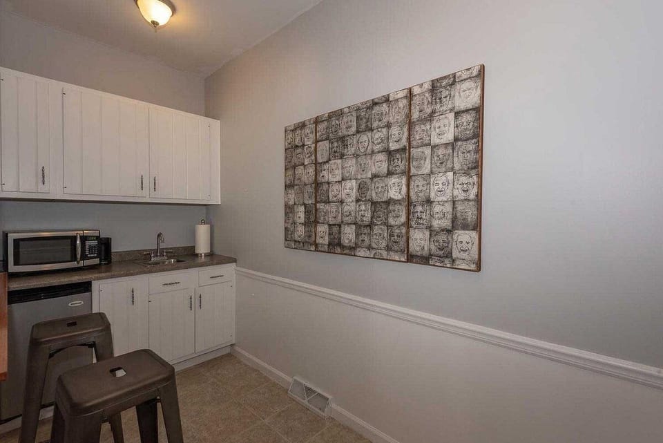 Compact kitchenette with white cabinets, dark countertop, stainless steel sink, microwave, and metal stools.