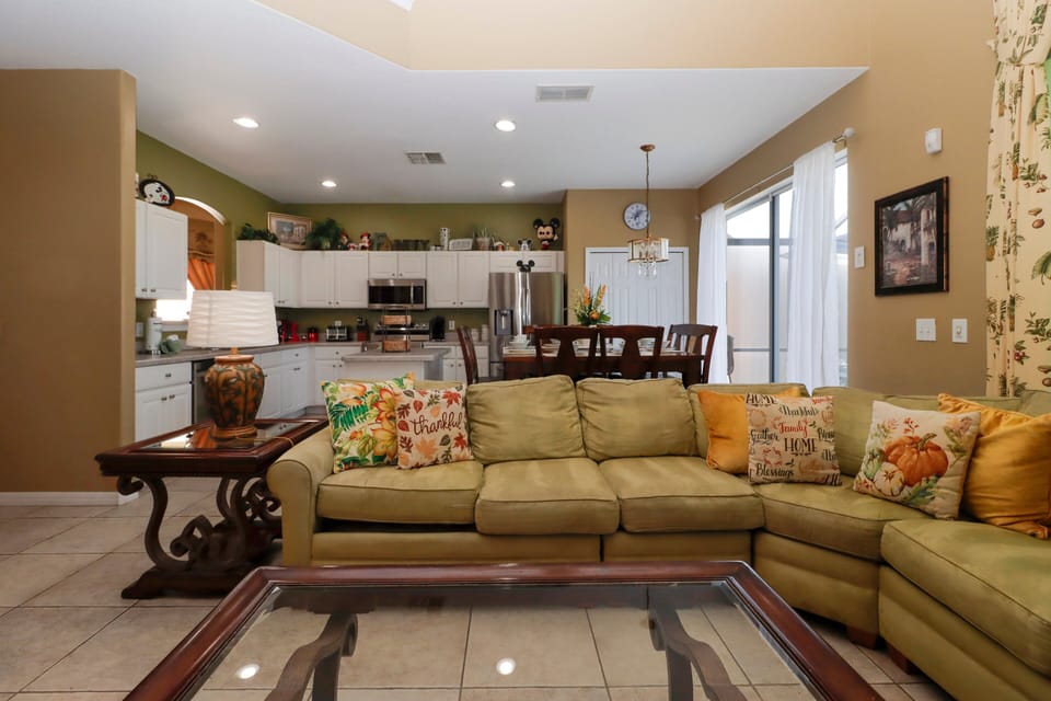 This spacious open-concept living area features a large L-shaped sectional sofa, flat-screen TV with entertainment console, two ornate end tables with lamps, a glass-top coffee table, tiled floors, vaulted ceilings.