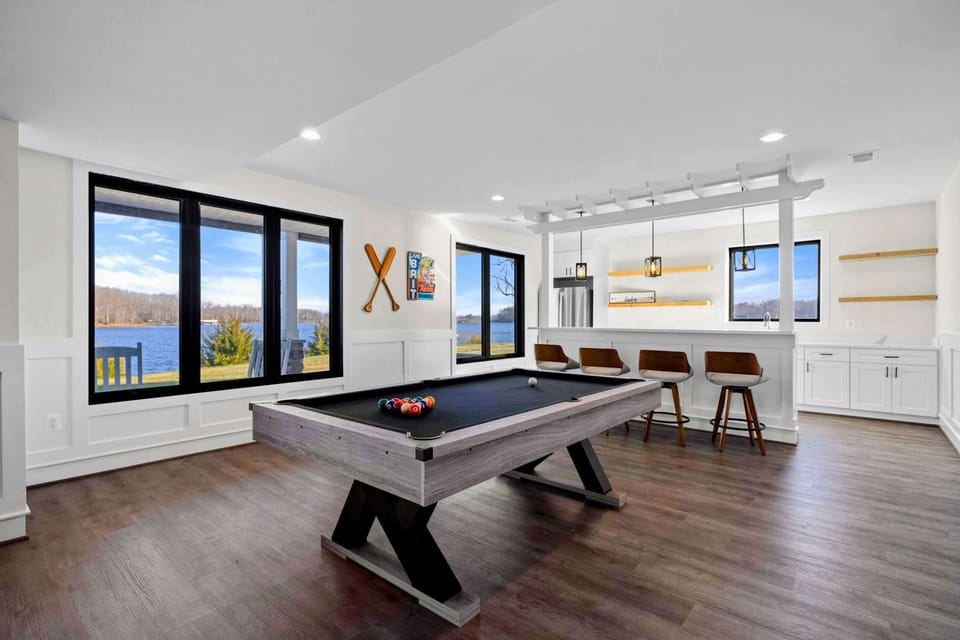 This spacious game room features a comfortable seating area with a modern fireplace, a pool table, ping-pong table, foosball table, shuffleboard, and a well-equipped kitchenette, all with stunning lake views.