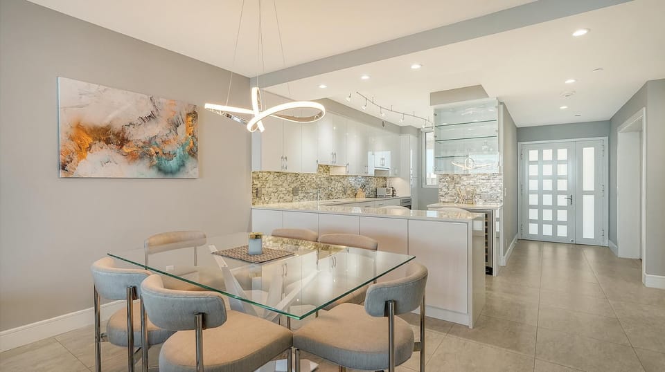 Dining area features a glass-top table with six cushioned gray chairs beneath a modern looped chandelier.