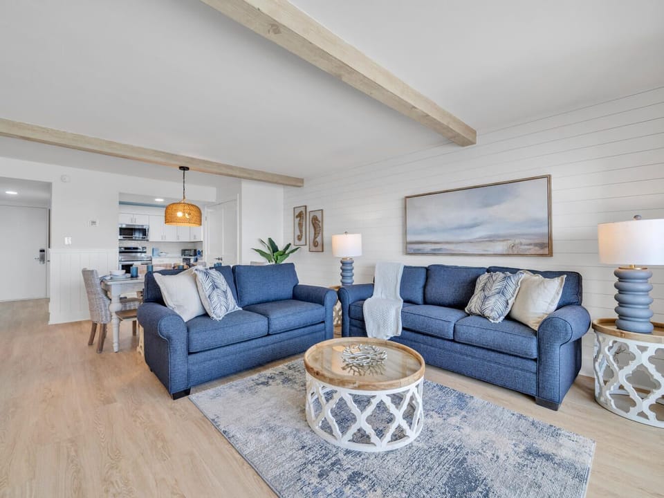 Relax in your coastal-inspired living space with plush blue sofas and warm wood beam ceilings creating the perfect atmosphere for unwinding.