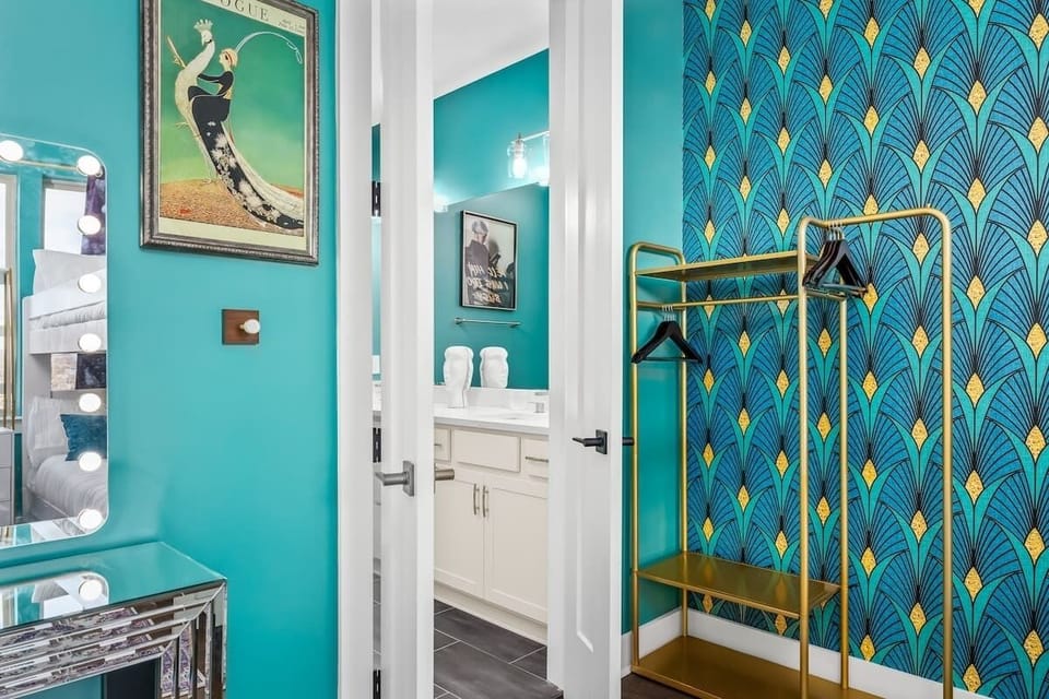 Sleep in style! This bold bedroom features teal walls, glam Art Deco wallpaper, a gold clothing rack, and a lit vanity perfect for getting ready. A chic, photo-worthy space designed for comfort and flair.