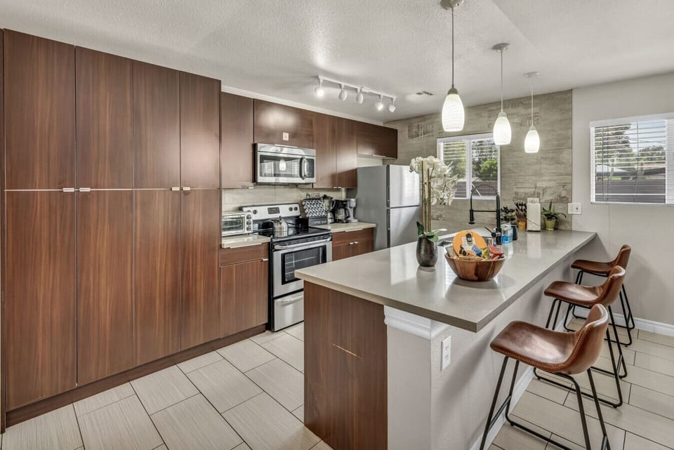 The kitchen includes brown cabinets, a Frigidaire microwave, Amana fridge, and Hamilton Beach oven. A Black+Decker toaster and coffee maker, plus a Hamilton Beach blender, are on the counter. A sink sits below a window, with pendant lights overhead.
